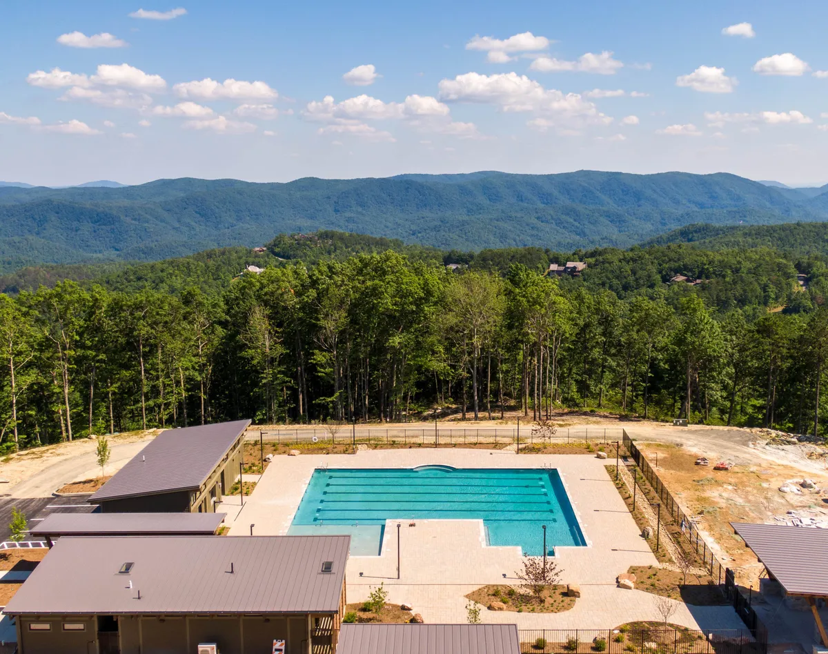 A beautiful exterior shot of a swimming pool surrounded by modern buildings and lush trees, with the impressive Blue Ridge Mountains rising in the background. The pool area is tranquil and well-maintained, providing a resort-like vibe ideal for wellness activities, relaxation, and connection with nature.