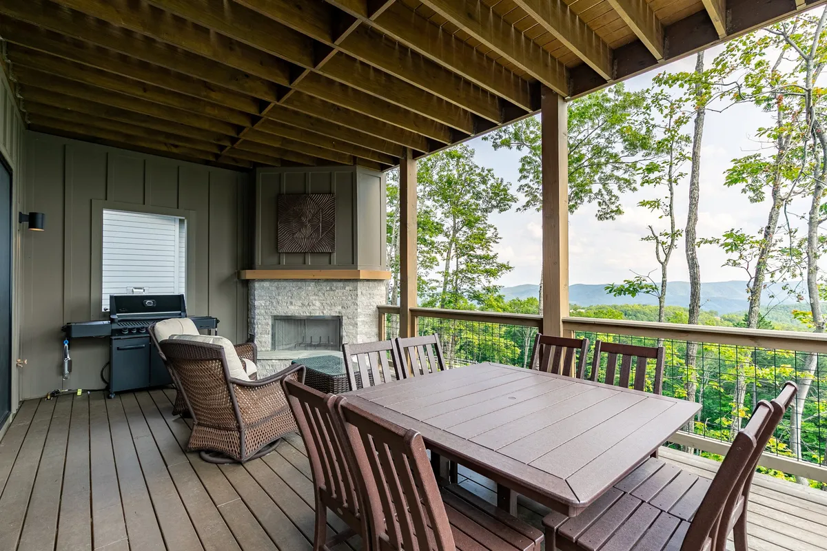 An expansive covered patio featuring a large wooden dining table set for eight, a stone fireplace, a built-in grill, and stunning views of surrounding trees and mountain peaks. The scene is bathed in warm sunset light, capturing the ambiance of outdoor living and dining.