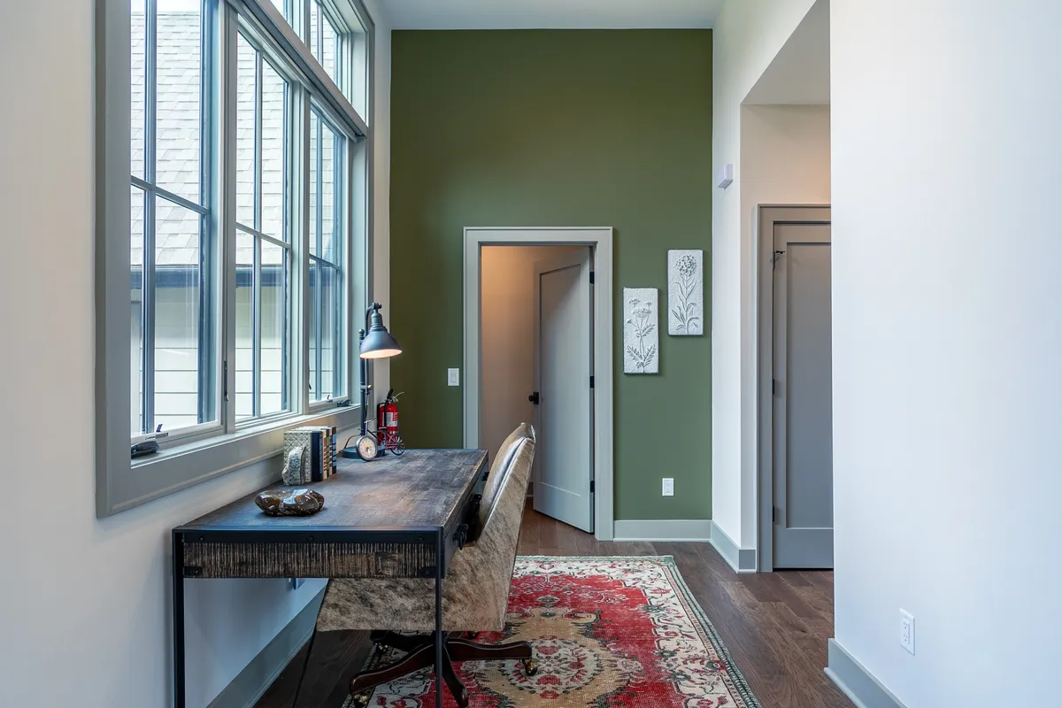 This image showcases a modern loft workspace with a desk and lamp set against a green accent wall, complemented by large windows that flood the area with natural light. Decorative artwork and serene décor create an inviting environment for work or study.