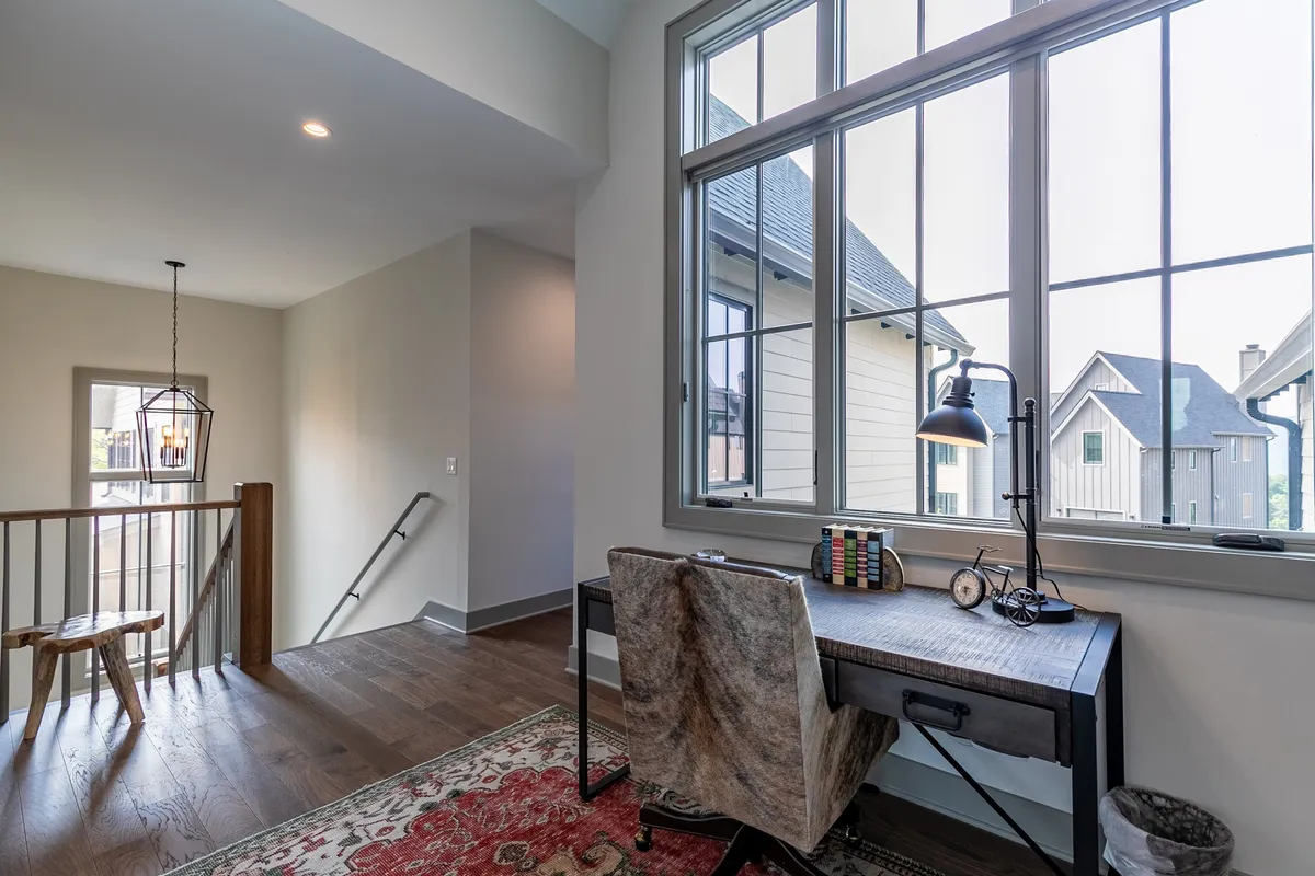 A peaceful workspace with a writing desk positioned near large windows, overlooking the outdoors. Soft natural light and a decorative rug contribute to a productive yet tranquil atmosphere ideal for focused work or study.