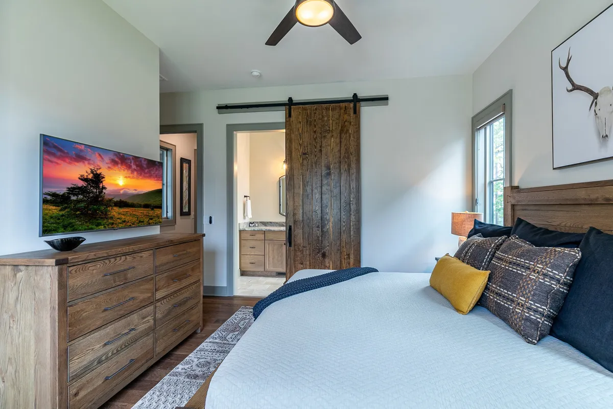 A cozy bedroom with a large bed, a dresser, a TV mounted on the wall, and a sliding barn door. Soft lighting and decorative artwork create a serene environment, while the windows offer hints of scenic views.