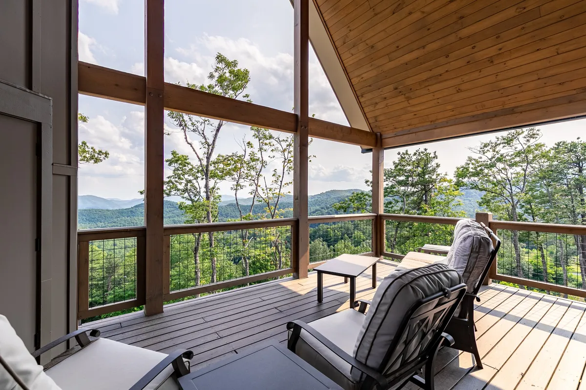 A comfortable seating area under a covered deck, with cozy furnishings and sweeping views of mountain ridges. The wooden ceiling and soft lighting evoke a warm, inviting setting perfect for morning coffee or evening lounging.