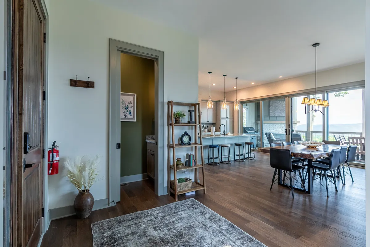 This image presents an inviting, open-concept interior featuring a dining table, an open kitchen, and a sitting area with large windows. Natural light bathes the space, and the arrangement seamlessly connects living, dining, and kitchen areas, offering a modern and sociable environment. The window frames beautiful mountain views, reinforcing the connection between indoors and outdoors.