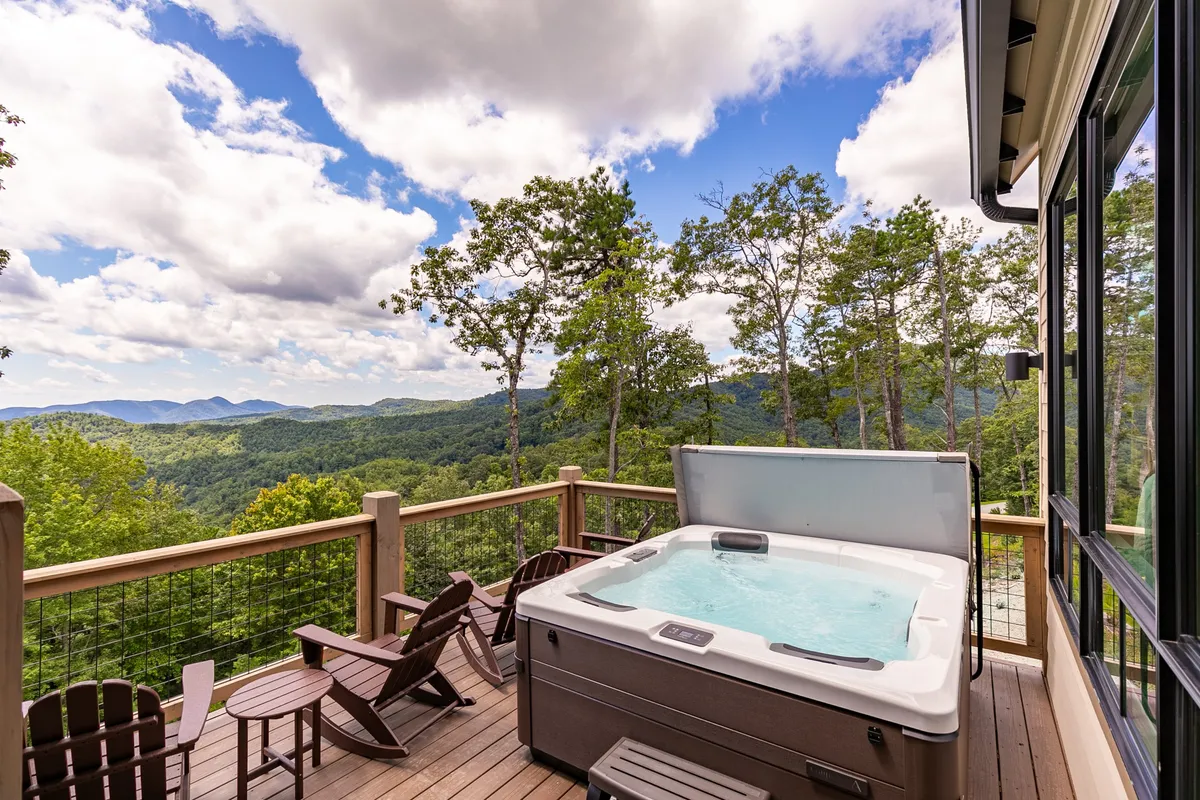 A serene exterior scene featuring a hot tub on a spacious deck, overlooking a sweeping mountain vista. Comfortable deck seating is arranged nearby, inviting guests to soak in the tranquil setting. The backdrop is filled with lush trees and rolling mountains, and the atmosphere is calm and rejuvenating, perfect for a wellness retreat.